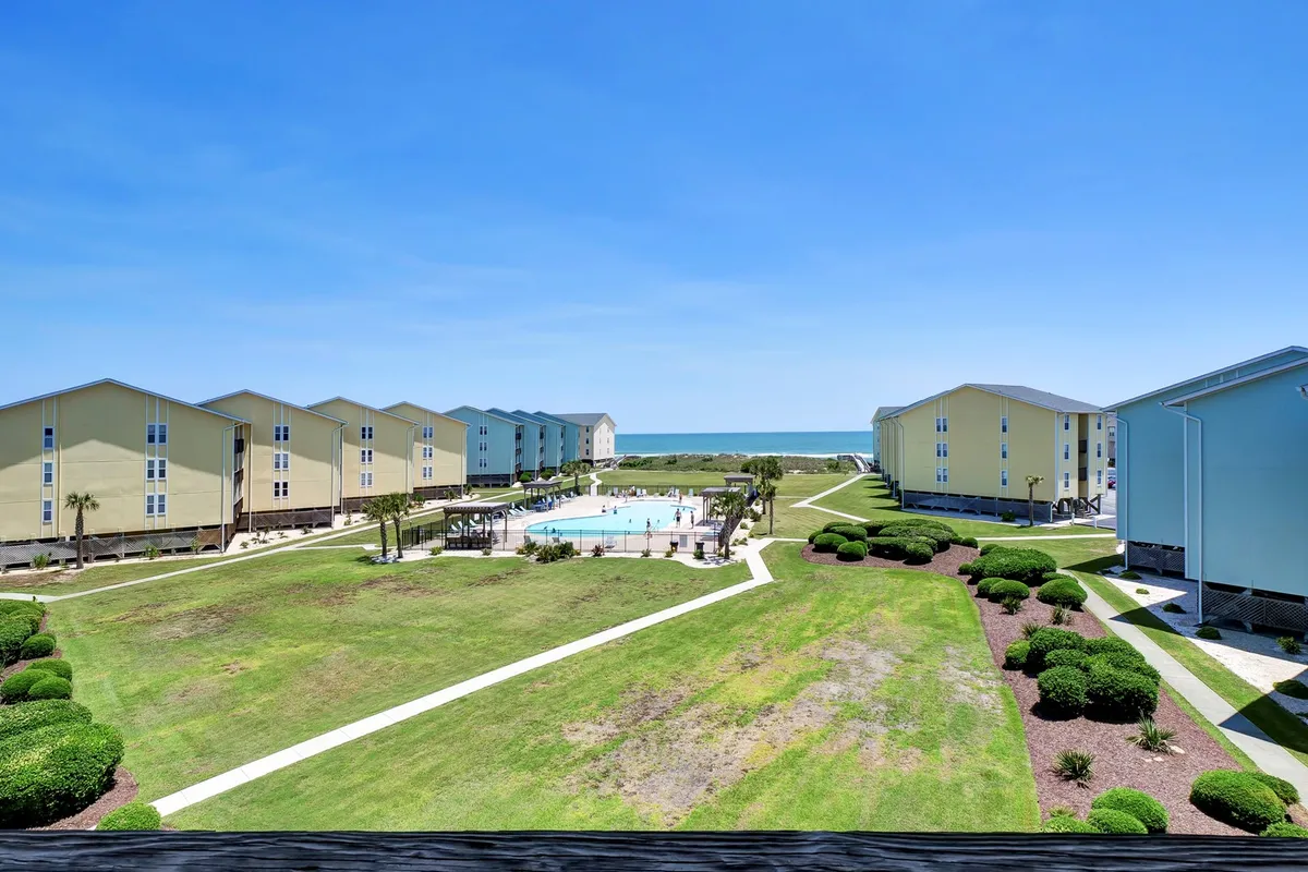 A vibrant exterior view featuring a resort-style swimming pool surrounded by landscaped lawns and with buildings and an ocean view in the background. The scene is bright, showing several loungers on the pool deck, hinting at a relaxed, communal environment.
