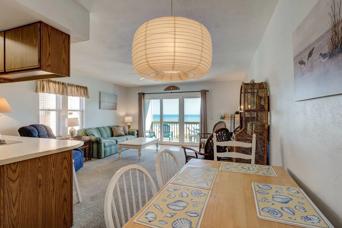 A serene interior scene with a large window offering a stunning ocean view, a dining table set with shell-patterned placemats, and a comfortable couch. The space is filled with natural light and decorated in calming coastal tones, inviting relaxation and social gatherings.
