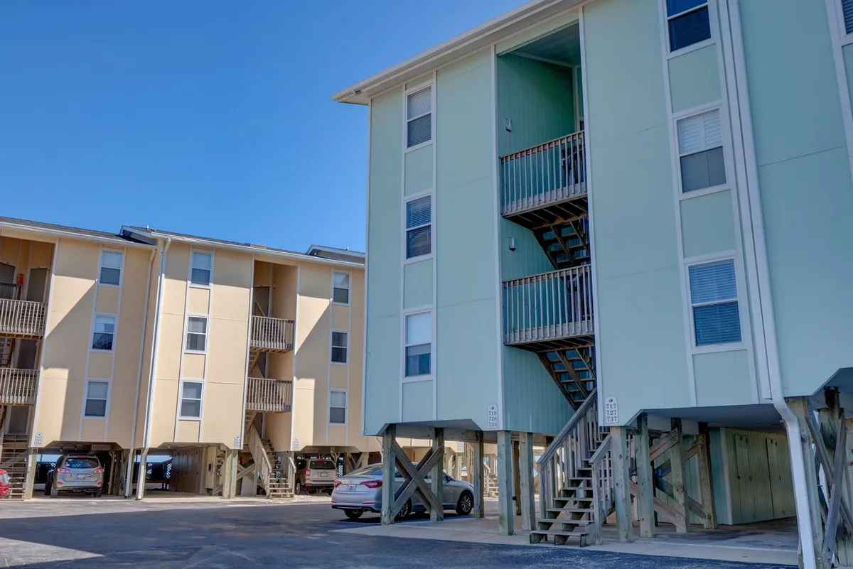 This image shows two multi-story buildings with a staircase leading up to upper levels. There are several cars parked underneath, suggesting ample parking and a welcoming, lively atmosphere. The scene exudes a casual and relaxed mood, with visible balconies ideal for gathering and enjoying sunset views.