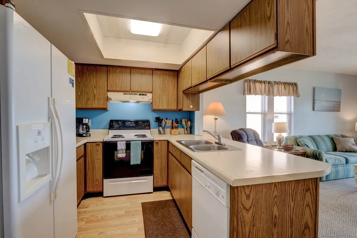 A warm, welcoming kitchen centered around a spacious island, complete with a stove, dishwasher, and sink. The open sightlines to the living area create a sense of togetherness, while the overall décor features subtle coastal touches. The island is ideal for meal prep, casual breakfasts, or evening entertaining.