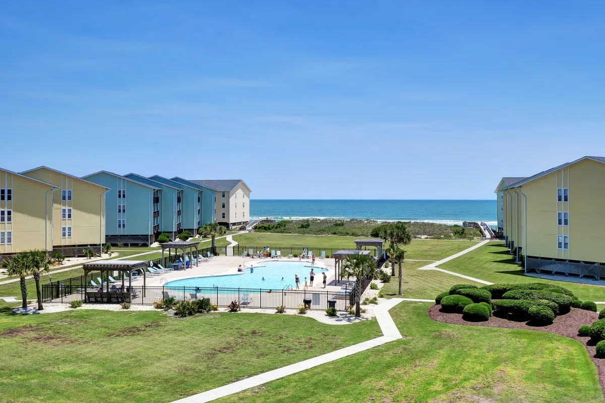 A wide shot of a communal swimming pool with sun loungers, a sandy beach just beyond, and apartment buildings in the background. Guests appear to be enjoying the pool and heading toward the beach, capturing the seamless connection between amenities and private beach access.