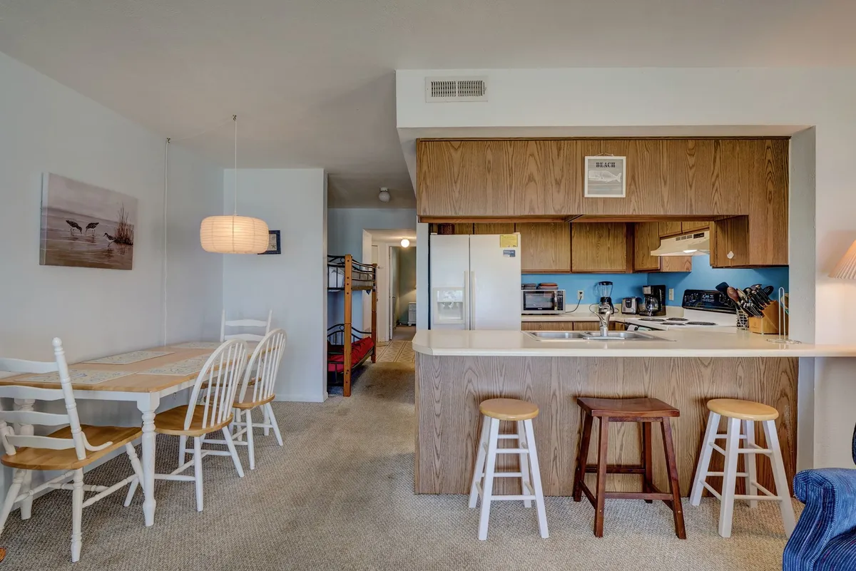 A casual, friendly dining and kitchen area featuring a kitchen island, a dining table set for meals, and nearby bunk beds that hint at a family-friendly atmosphere. The light fixture and layout create an inviting space for sharing meals or playing games after a day at the beach.