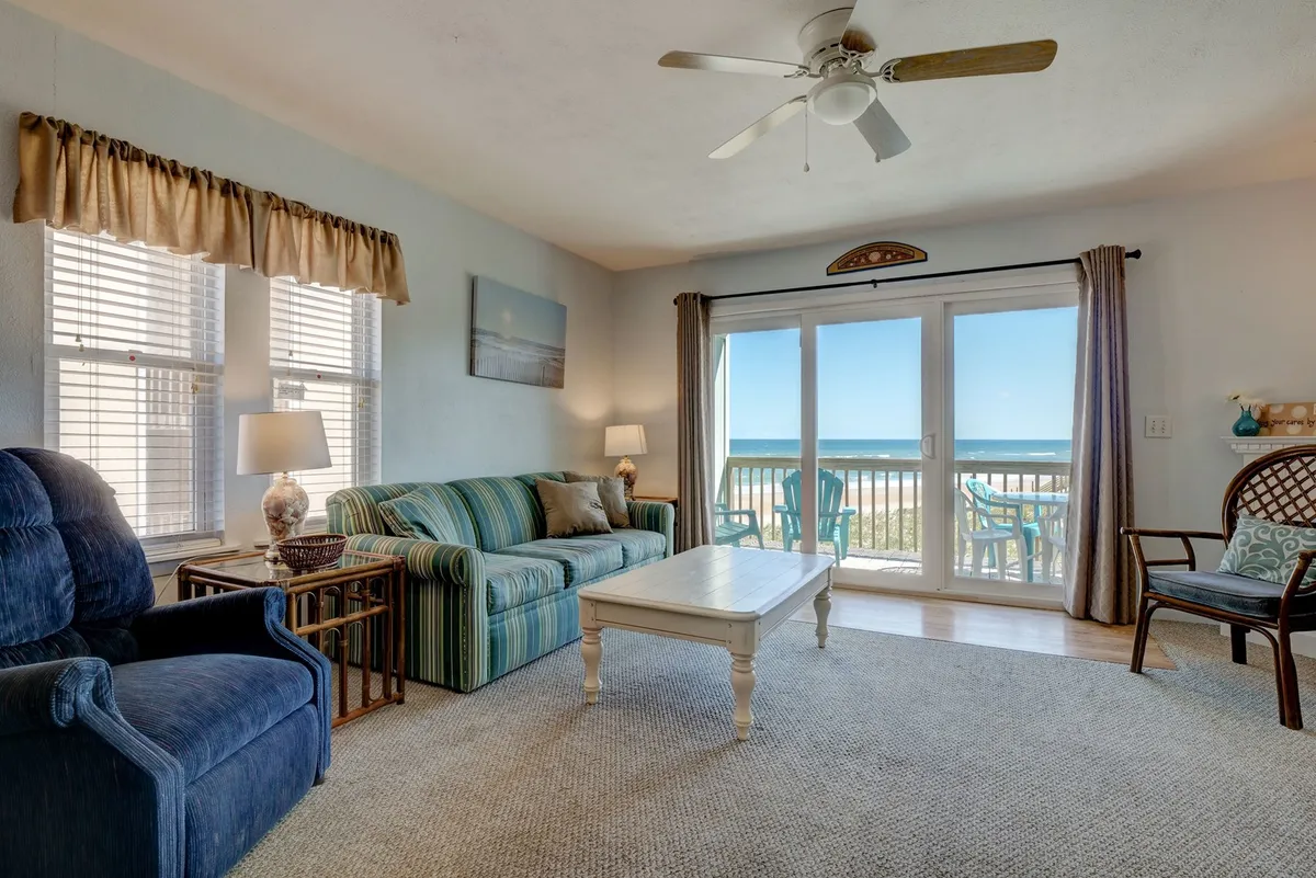 This image captures a spacious living room featuring a large sliding glass door that frames a direct ocean view. A striped couch and coffee table create a cozy sitting area, bathed in natural light and coastal hues, while the glass doors hint at seamless access to a private deck beyond.
