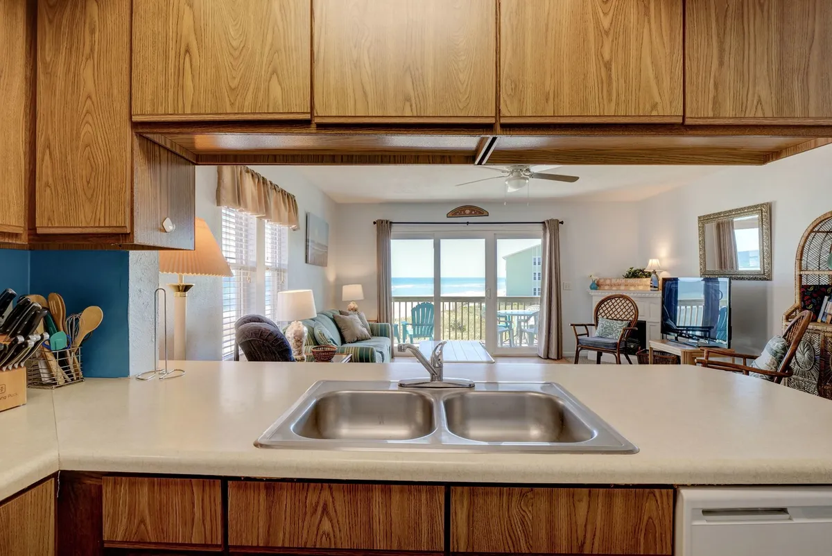 A bright and airy kitchen featuring wooden cabinetry, a spacious kitchen sink, and a large window that lets in plenty of natural light. The open plan layout allows for a seamless flow between the kitchen and the living area, with space for cooking, socializing, and relaxing. The window provides a scenic view, enhancing the coastal ambiance.