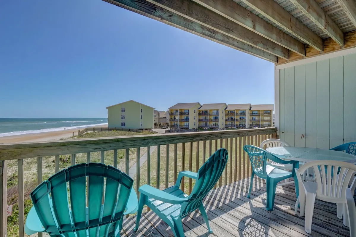 The image showcases a second-floor deck with turquoise Adirondack chairs and an outdoor dining table, all overlooking stunning ocean views and beachfront condo buildings. The inviting setup suggests a perfect spot for relaxing, dining, and socializing while surrounded by the serene coastal landscape.