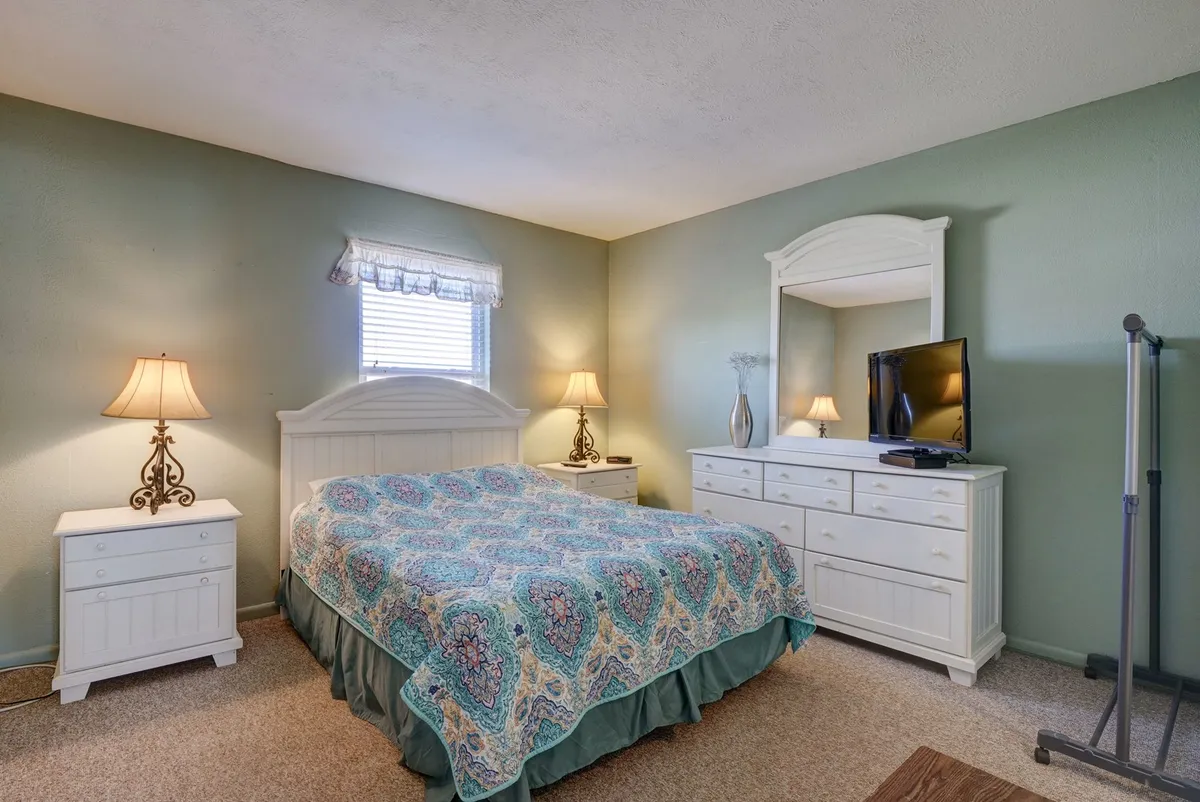 A bright and inviting master bedroom, featuring a queen-sized bed with patterned bedding. White furniture, including bedside tables with lamps and a dresser with a mirror, create a cozy and comfortable atmosphere. The space feels restful and perfect for relaxing or enjoying a quiet evening, and a television is visible for entertainment.